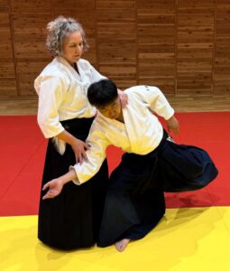 Senior aikido instructor in a white gi assists a student in a white gi and black hakama during a throw on a red-yellow mat.